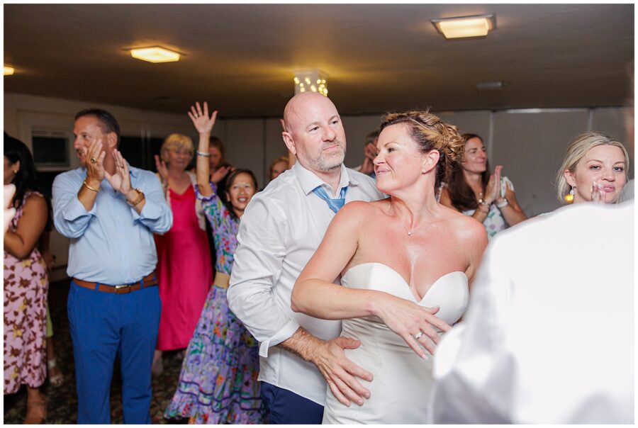 Bride and groom embrace on the dance floor surrounded by cheering guests, sharing a playful and romantic moment during their Cape Cod wedding reception, representing the portfolio of a Cape Cod wedding photographer.