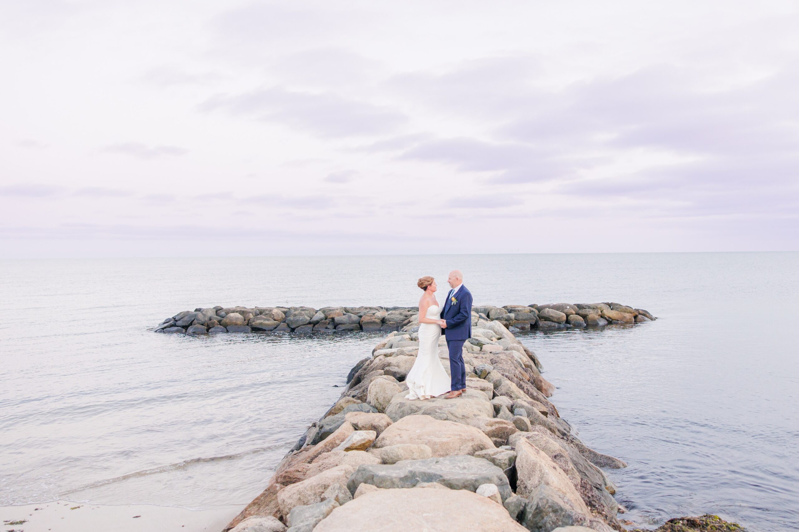 Bride and groom standing together on a rock jetty surrounded by ocean waves. A cinematic Cape Cod waterfront wedding portrait.