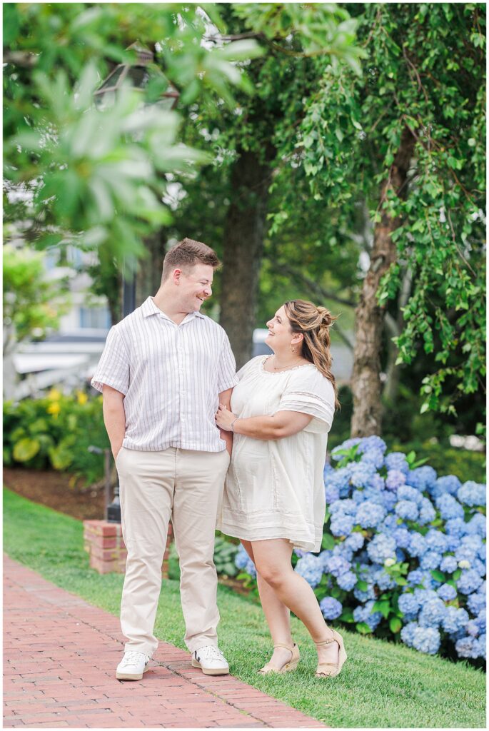 Couple standing on a brick walkway at Chatham Bars Inn during summer engagement session, surrounded by lush green trees and blooming blue hydrangeas
