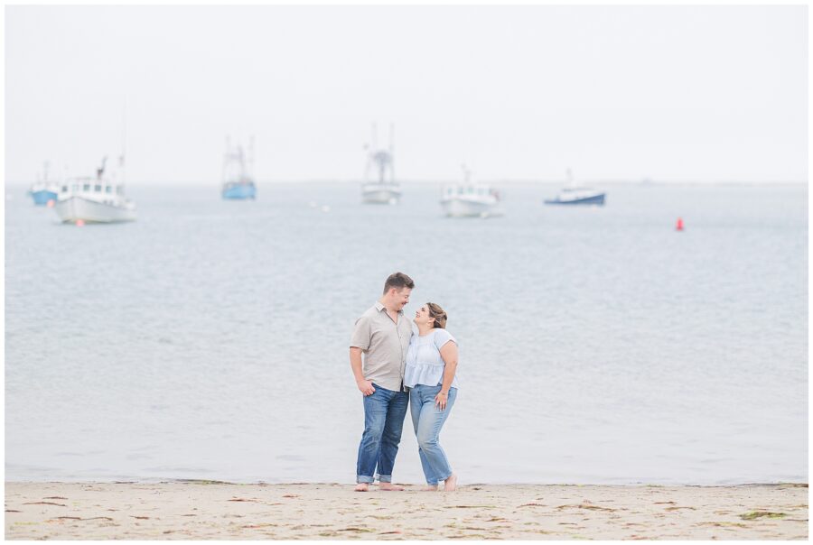 Engaged couple barefoot on the private beach at Chatham Bars Inn, standing close with Cape Cod fishing boats in the background