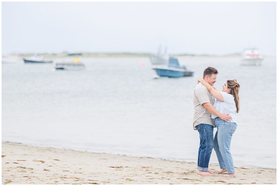 Groom-to-be hugging bride-to-be on Chatham Bars Inn’s private beach with boats in the distance
