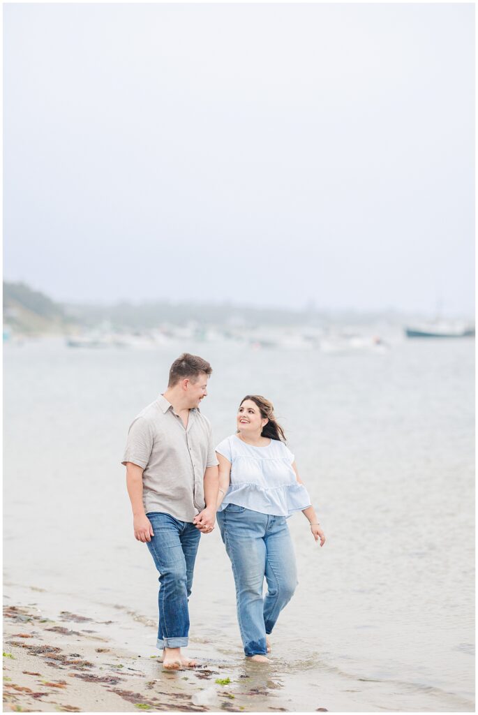 Engaged couple walking barefoot along the shoreline at Chatham Bars Inn with Cape Cod boats in the background