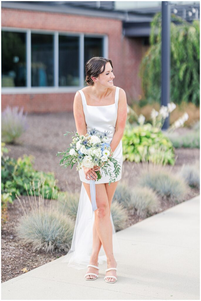 Bride smiling in a short white dress holding a bouquet of white and blue flowers, photographed by a Boston Elopement Photographer.