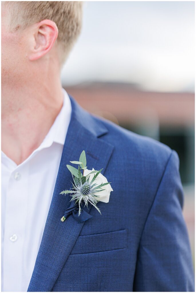 Close-up of the groom’s boutonniere with a white rose and thistle pinned to his blue suit.
