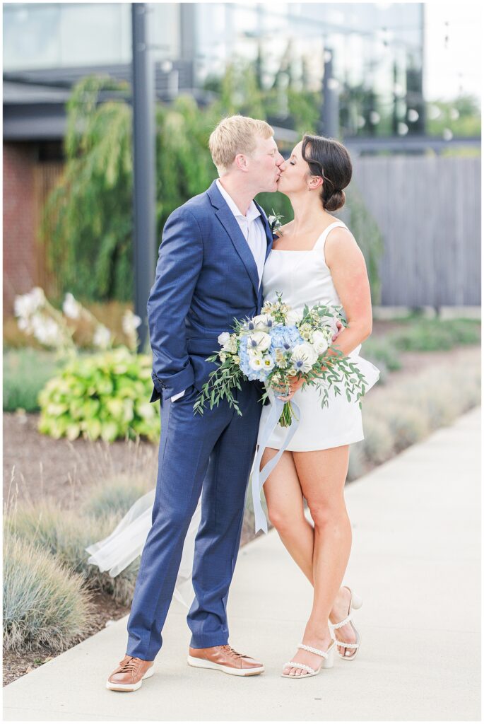 Bride and groom share a kiss outdoors, the bride holding a bouquet of white and blue flowers, captured by a Boston Elopement Photographer.