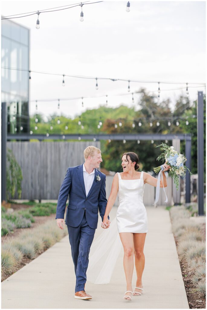 Couple holding hands and walking under string lights, the bride raising her bouquet with joy during their Boston elopement.