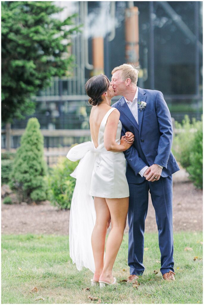Bride and groom kissing while standing on grass with a modern brewery backdrop.