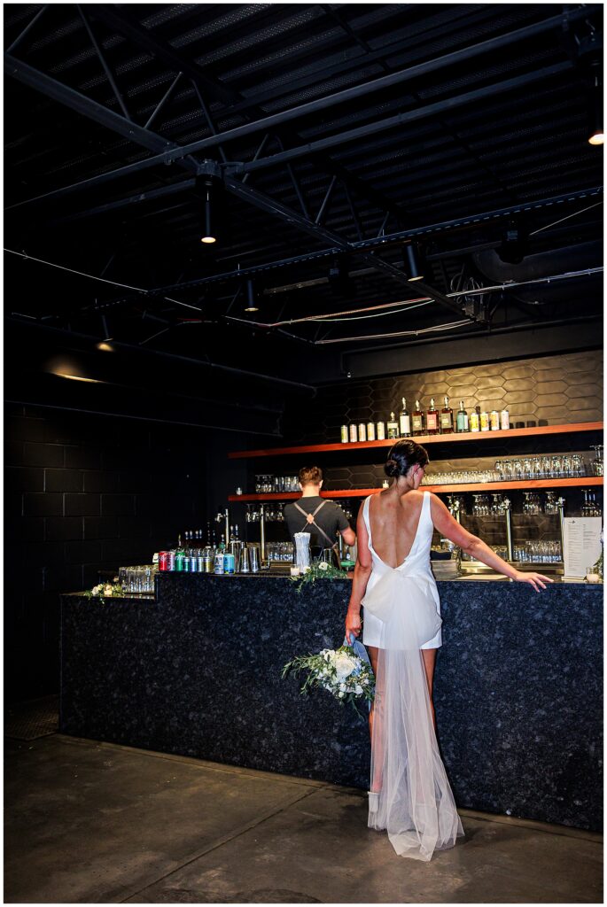 Bride in a short white dress with a long train stands at a bar inside Trillium Brewery, holding her bouquet.
