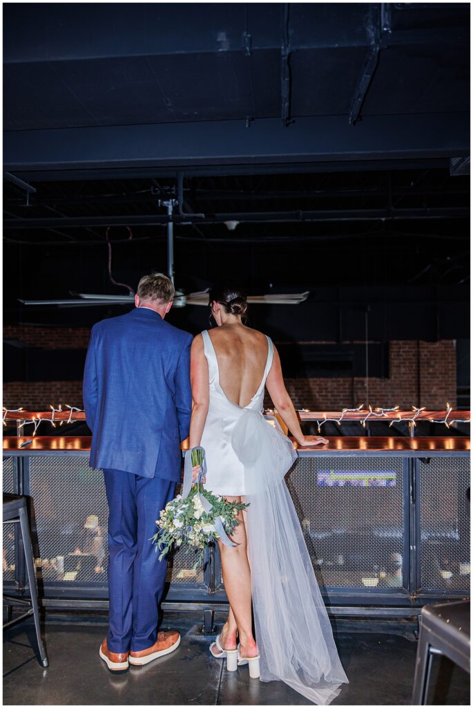 Bride and groom stand together indoors, looking out from a balcony decorated with string lights, captured by a Boston Elopement Photographer.