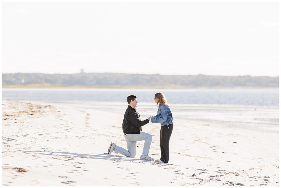 A man kneels on one knee in the sand, holding hands with a woman on a quiet beach. The ocean stretches behind them under soft sunlight.