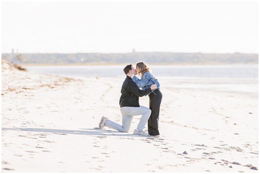 The same couple kisses as the man remains kneeling on one knee in the sand, with the shoreline and calm waves in the background.