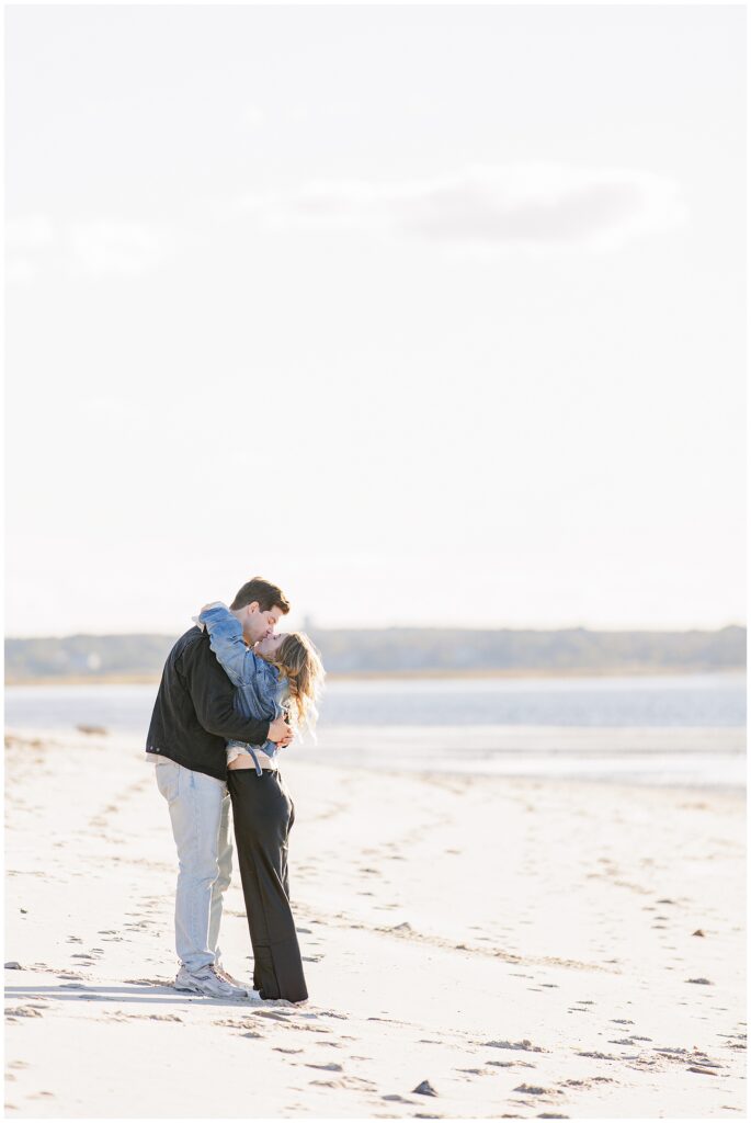 The couple stands embracing on the beach, holding each other close near the water’s edge with a distant shoreline in the background.