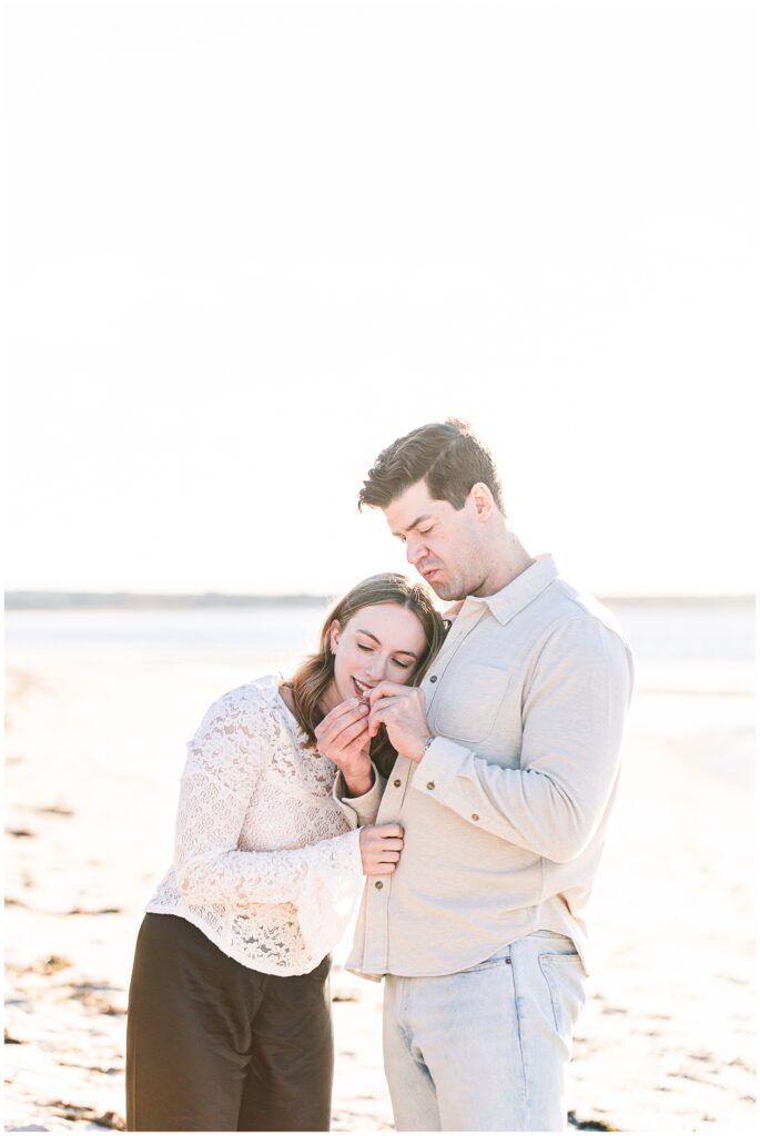 The couple stands close together on the beach, looking at a ring in the man’s hand as the woman leans her head on his shoulder.