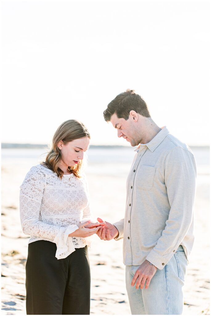 The couple stands facing each other, both looking down at the woman’s hand as she admires a ring. The ocean is softly blurred in the background.