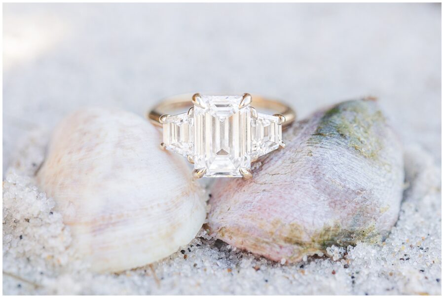 A close-up of a gold engagement ring with a large emerald-cut diamond flanked by two smaller diamonds, resting on seashells in the sand.