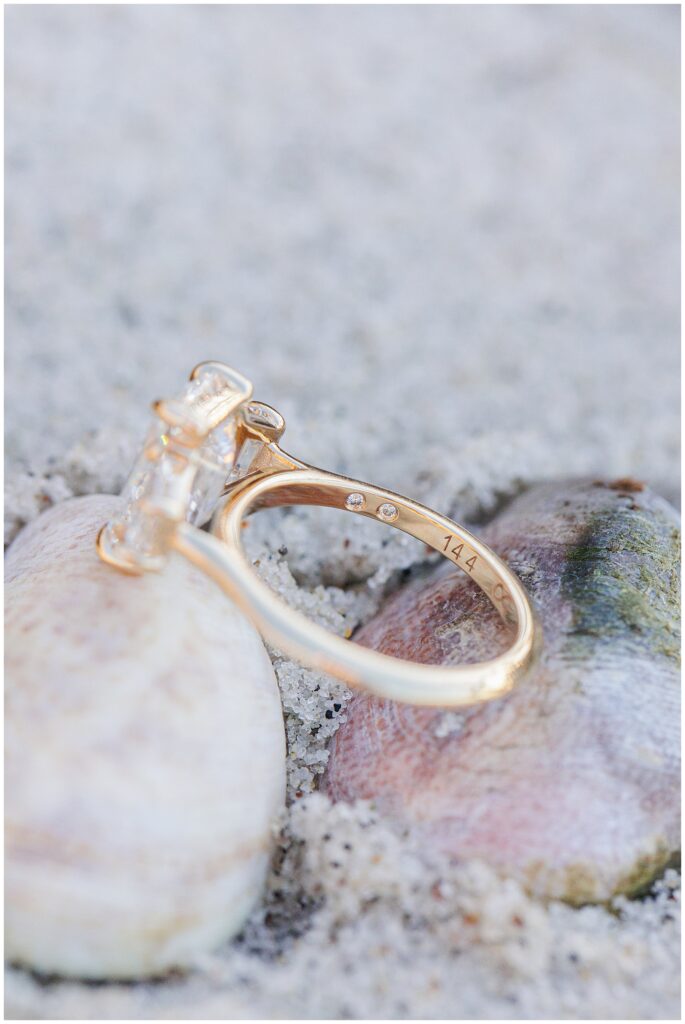 A side view of the same gold engagement ring resting on seashells in the sand, showing small inset diamonds and an engraving on the band.