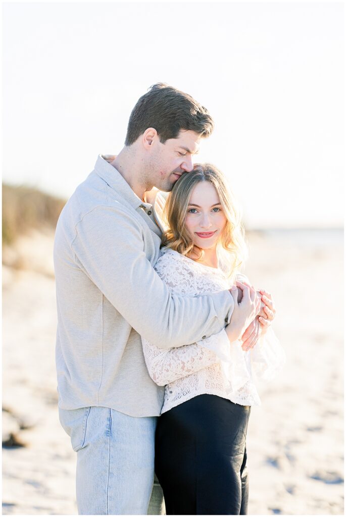 The couple embraces on the beach, with the man holding the woman from behind and resting his head against hers as she looks toward the camera.