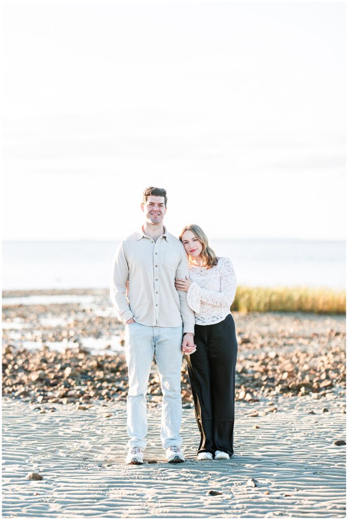 A couple stands on the sand at low tide, holding hands and smiling. The woman leans on the man’s shoulder with the ocean and rocks behind them.
