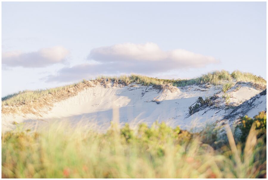A view of a large sand dune covered with grass, softly lit in the late-day sunlight beneath a few clouds.