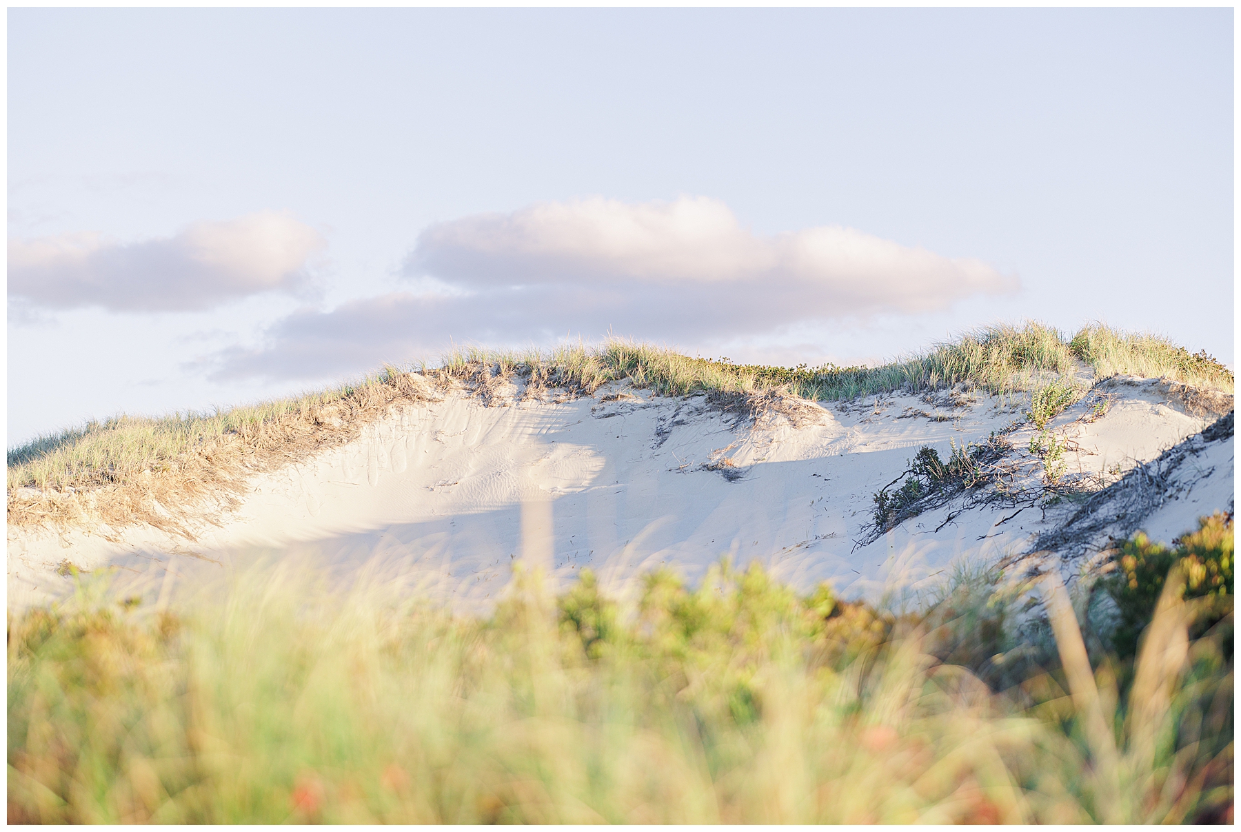 A view of a large sand dune covered with grass, softly lit in the late-day sunlight beneath a few clouds.
