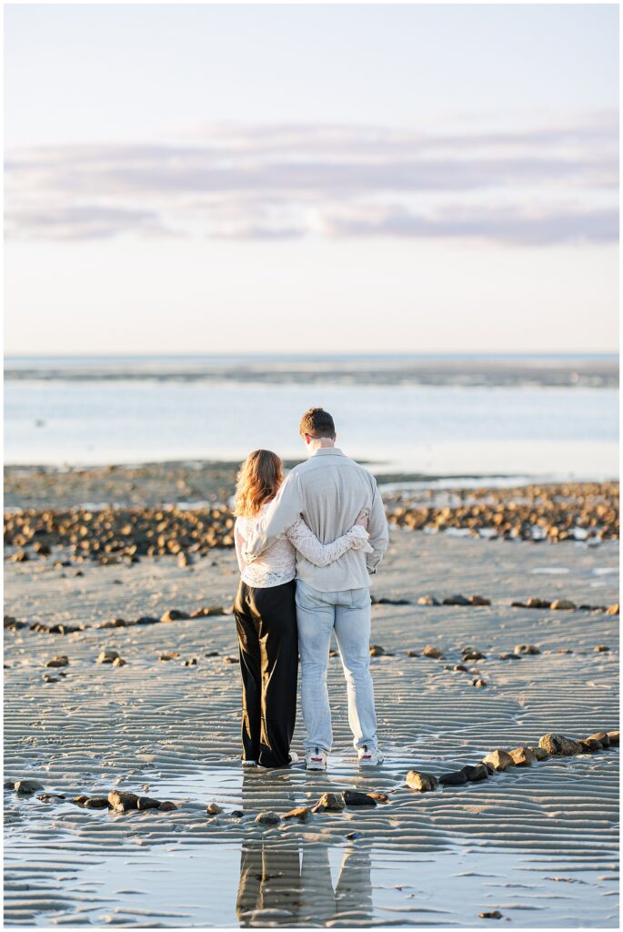 The couple stands arm in arm, facing the ocean at low tide. Ripples in the wet sand and scattered rocks reflect the golden light.