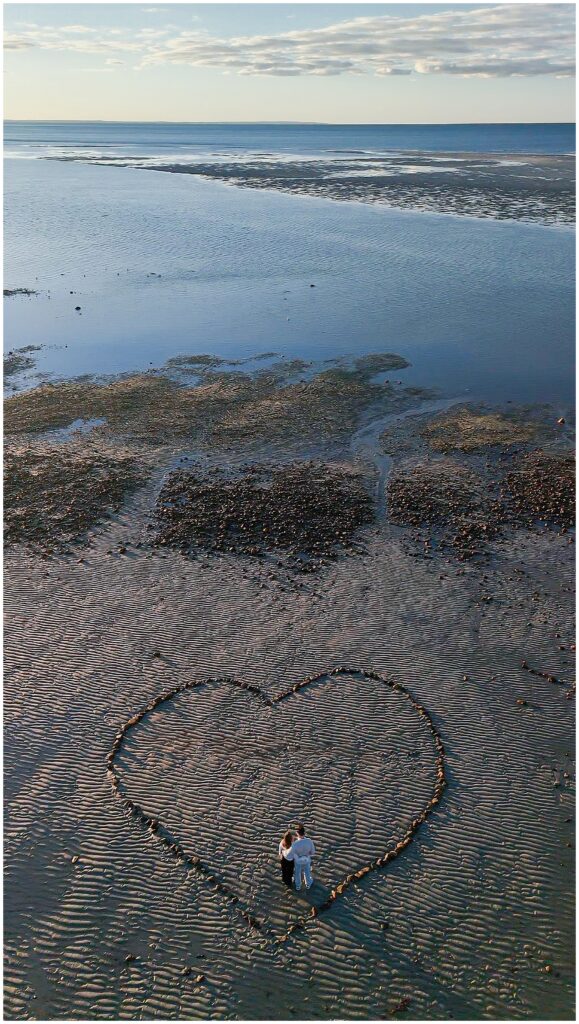 Aerial view of the couple standing inside a large heart shape outlined with rocks on the rippled sand at low tide.