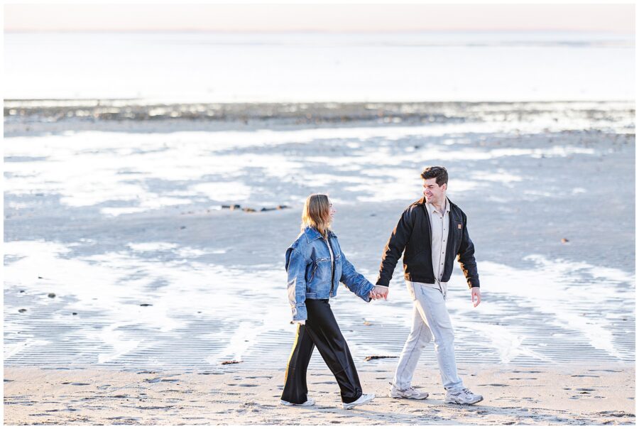 The couple walks hand in hand along the beach, smiling at each other with water shimmering in the background.
