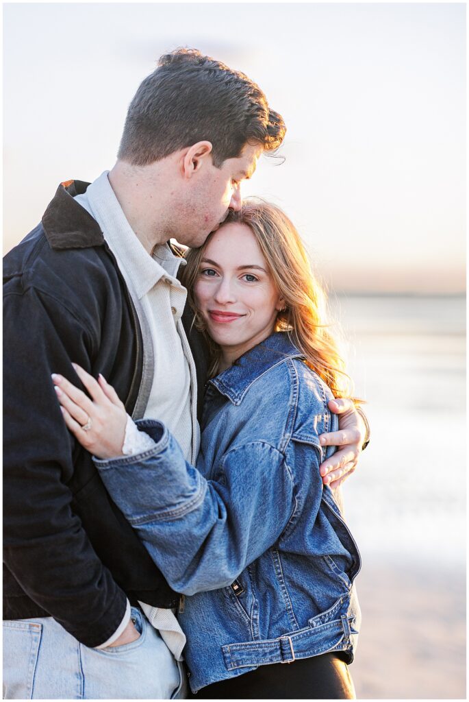 The man kisses the woman’s forehead as she looks at the camera, smiling gently while they embrace on the beach.