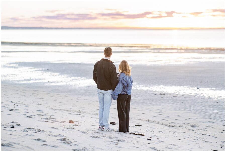  The couple stands side by side on the sand, facing the glowing sunset over the ocean horizon.