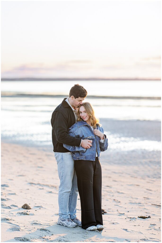 The couple embraces closely on the beach, with gentle evening light reflecting off the water behind them.