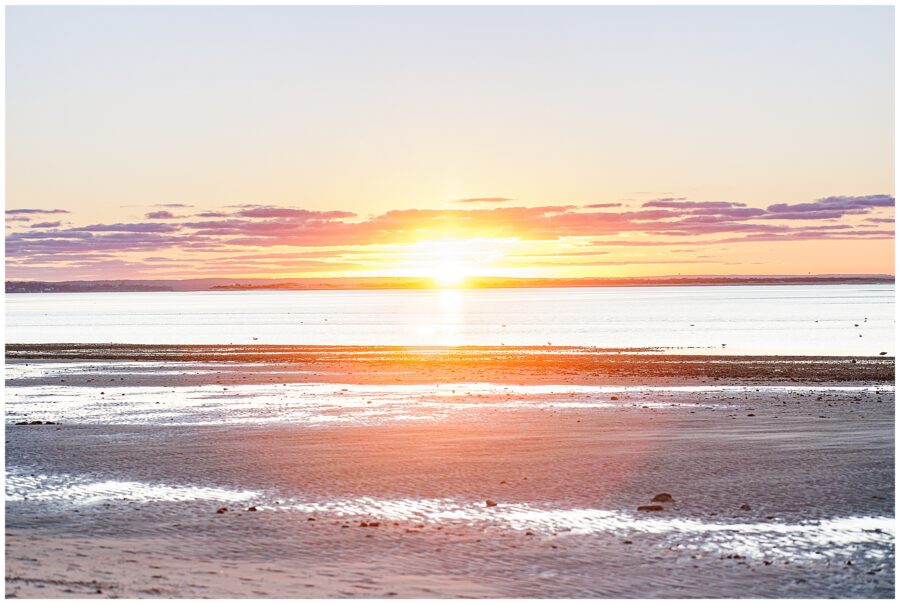 The sun sets over Chapin Beach, casting orange and pink light across the rippled sand and calm ocean.
