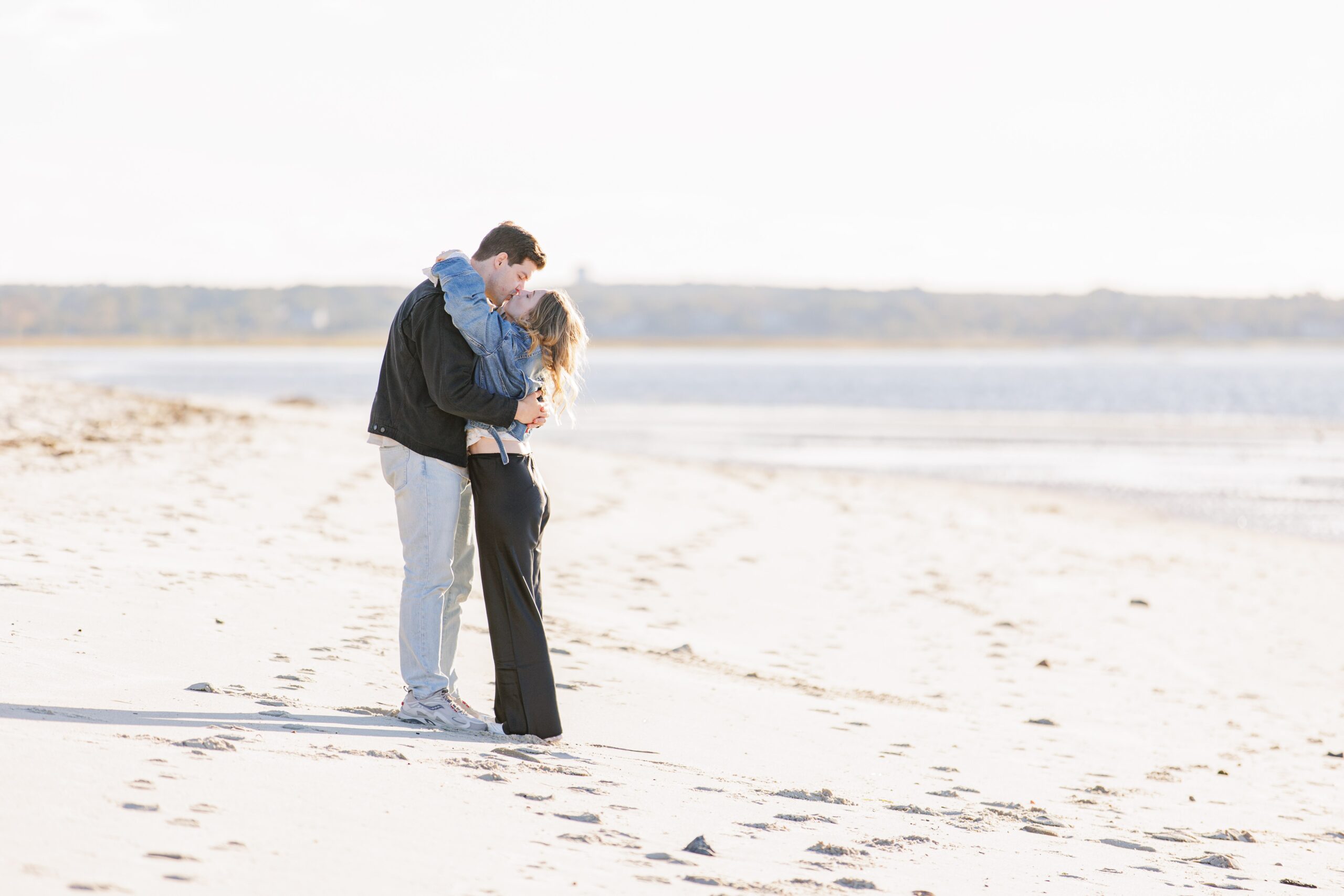 A man and woman stand on a sandy beach near the shoreline, embracing and kissing with the ocean and distant dunes behind them in soft sunlight.