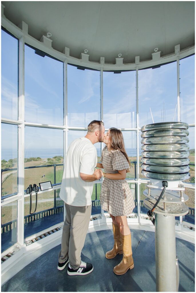 The couple kisses inside the lighthouse lantern room, holding hands with ocean views visible in the background.