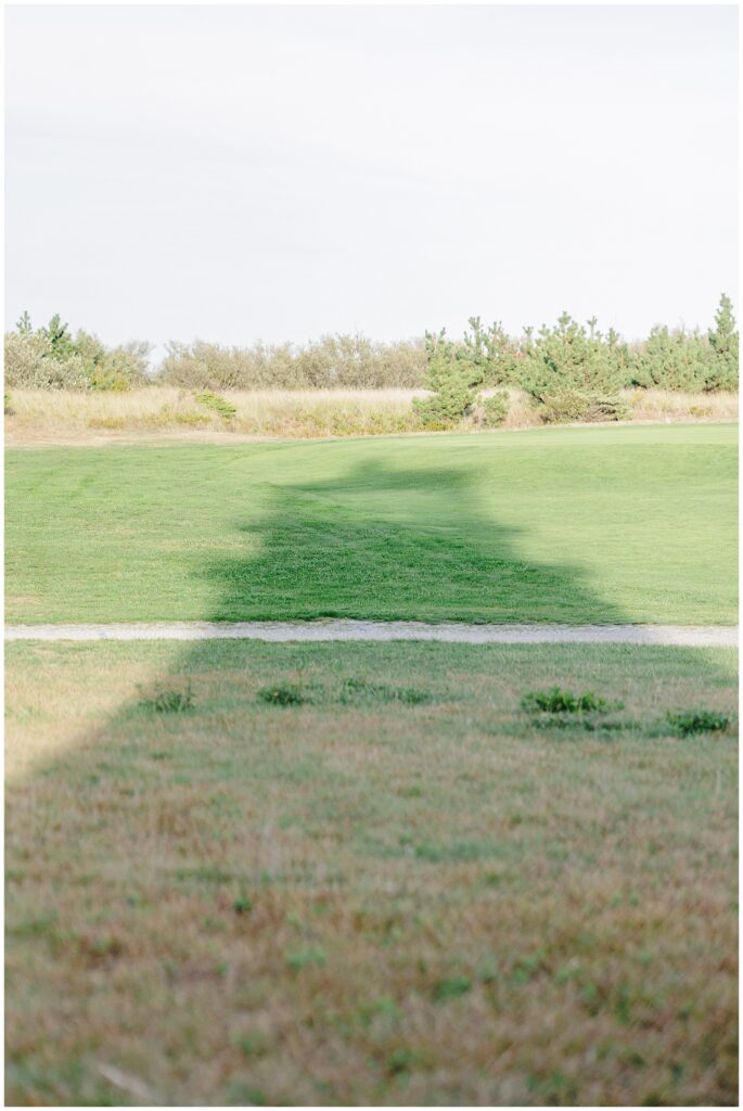 The shadow of Highland Lighthouse stretches across a grassy field bordered by low trees and shrubs.
