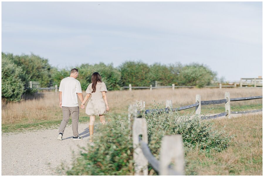 The couple walks hand in hand down a dirt path lined with wooden fences and tall grasses near Highland Lighthouse.