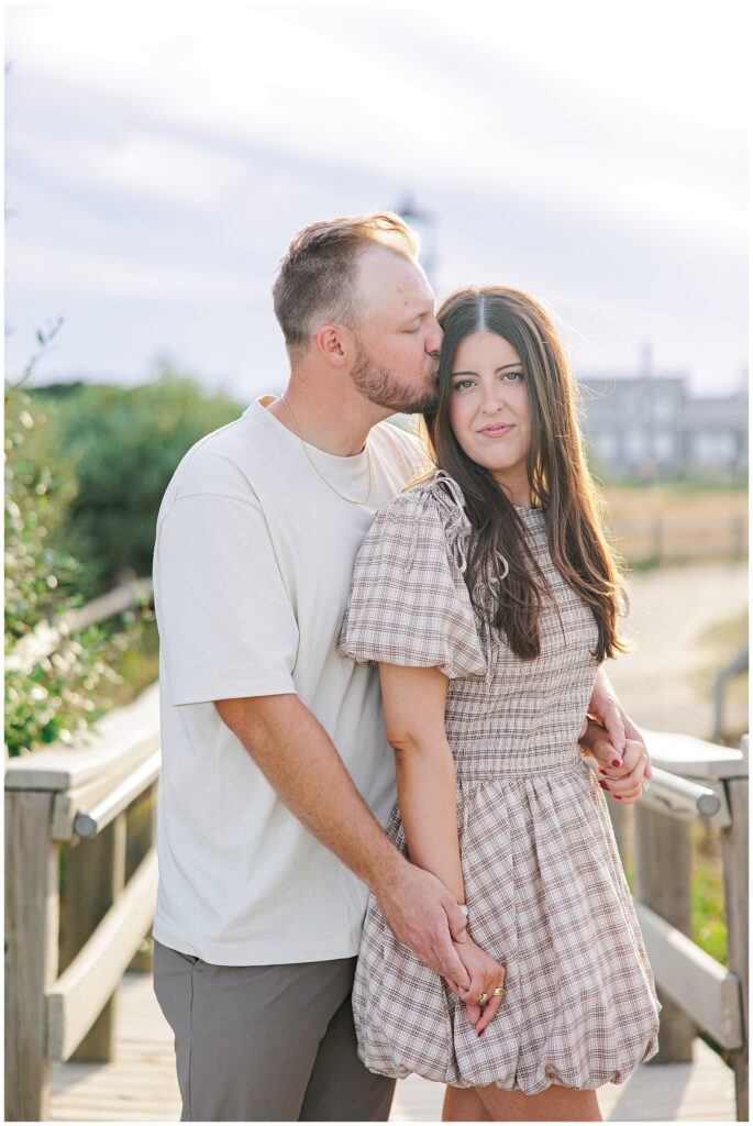 The man stands behind the woman on a wooden walkway, kissing her on the temple as she smiles slightly. The lighthouse and buildings appear blurred in the background.