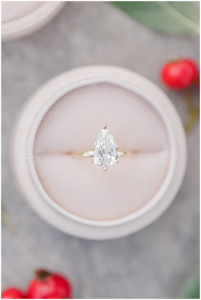 A close-up of a pear-shaped diamond engagement ring with a gold band, displayed in a light pink velvet ring box with red berries nearby.