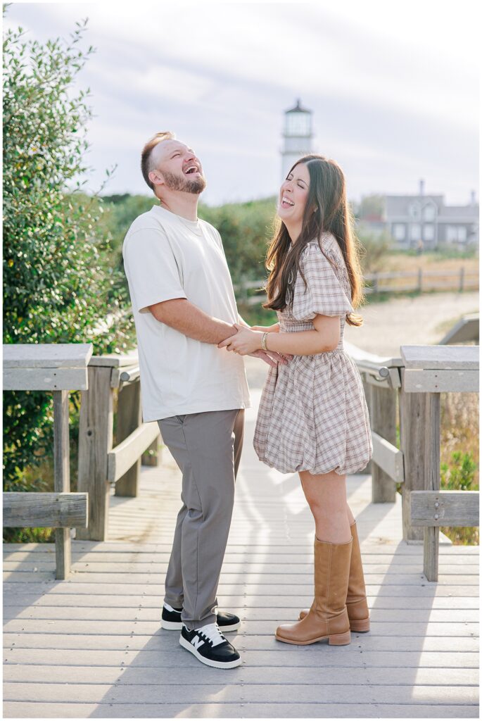 The couple stands laughing together on a wooden walkway, holding hands. The lighthouse is visible in the distance behind them.
