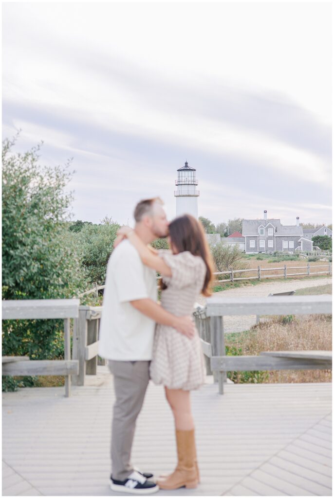 A couple embraces and kisses on a wooden deck with Highland Lighthouse and nearby buildings softly blurred in the background under a pastel sky.