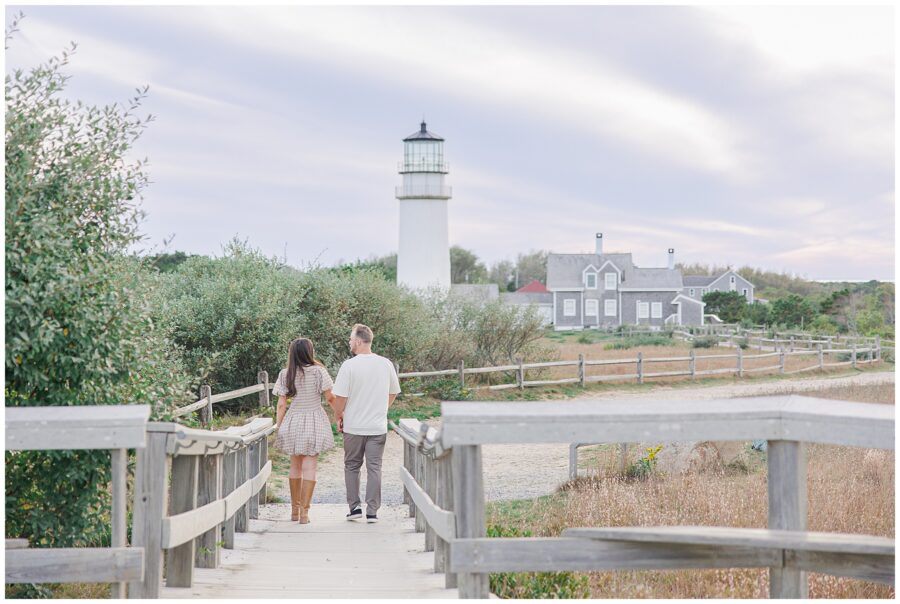 The couple walks hand in hand down a wooden walkway toward Highland Lighthouse, surrounded by greenery and weathered fences.