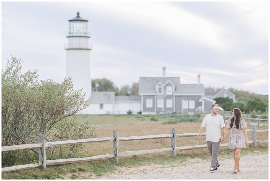 The couple walks along a dirt path beside a wooden fence, smiling at each other, with Highland Lighthouse and gray shingled keeper’s houses behind them.