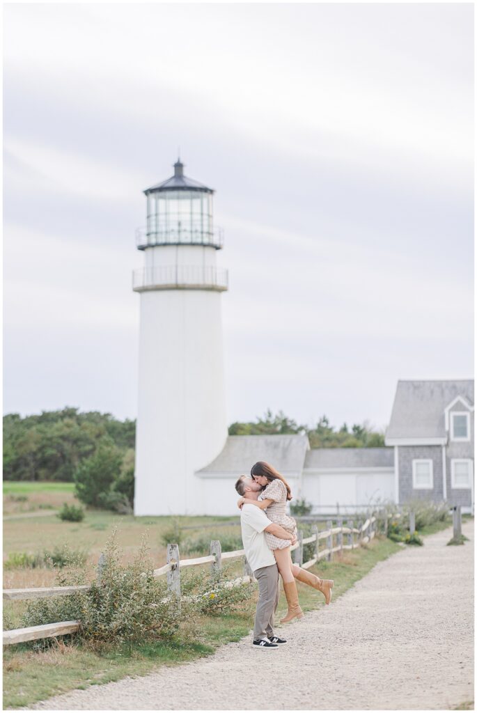 The man lifts the woman off the ground as they kiss beside a wooden fence, with Highland Lighthouse and its buildings in the background.