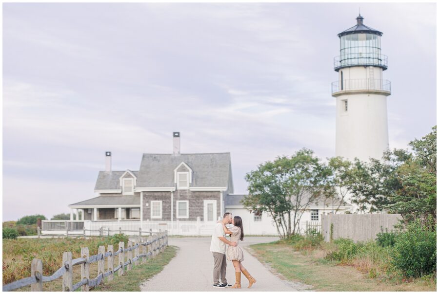 The couple kisses on a path leading to Highland Lighthouse, with the lighthouse and keeper’s house framed by a soft sky and greenery.