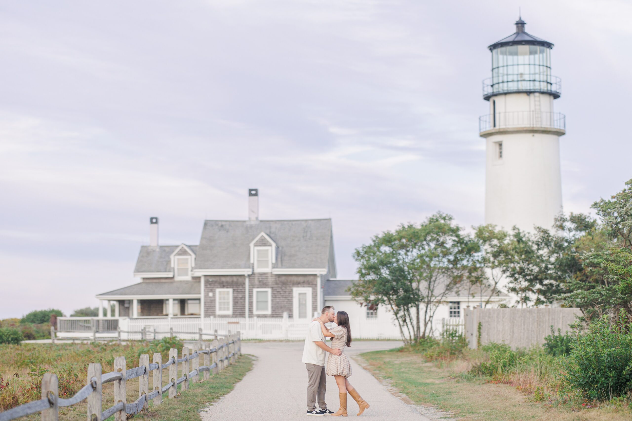 The couple kisses on a paved path in front of Highland Lighthouse and the gray shingled keeper’s house, with soft evening light in the sky.