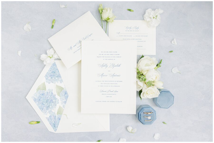 Flat lay of elegant wedding invitations and stationery on a light blue surface, accented with white flowers, blue velvet ring boxes, and gold rings. Invitation text includes “Ashley Elizabeth and Marco Antonio” with Nauset Beach Inn, Orleans, Massachusetts listed as the venue.