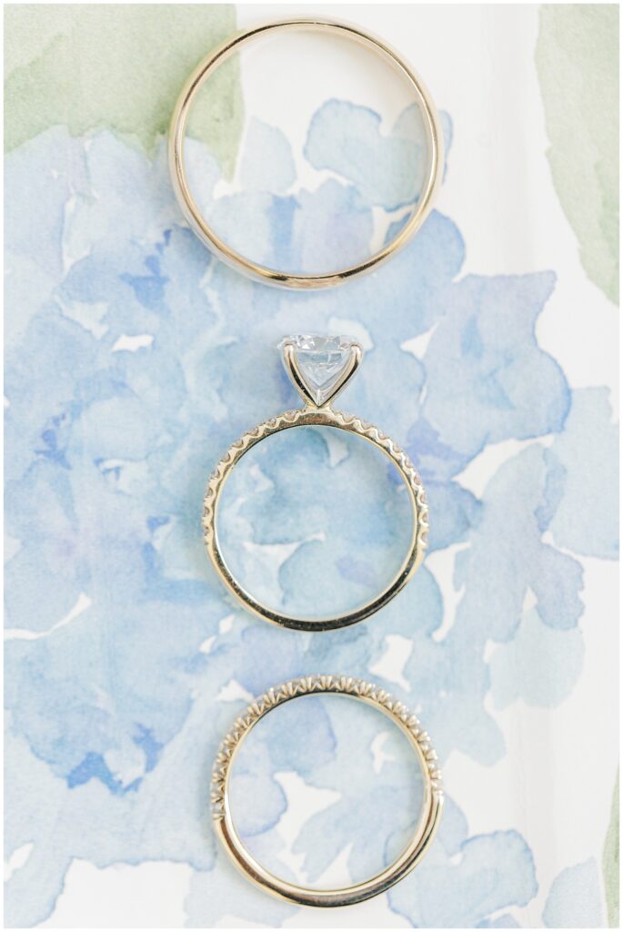 Close-up of three gold wedding rings arranged vertically on floral blue watercolor paper — a plain band, an engagement ring with a diamond solitaire, and a diamond-studded band.
