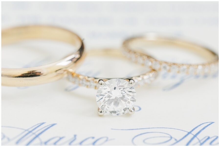 Detailed close-up of three gold rings, including a solitaire engagement ring with a round diamond and two matching gold wedding bands, resting on white paper with blue calligraphy text.