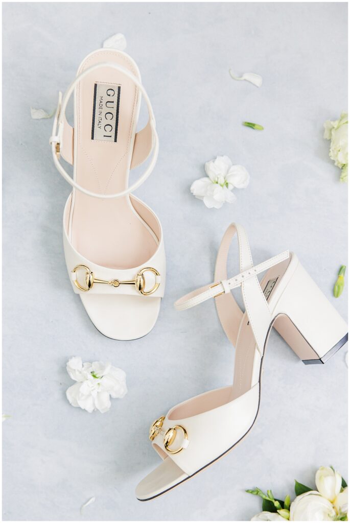 Pair of white Gucci block-heeled sandals with gold horsebit details, arranged on a pale blue background with scattered white flowers.