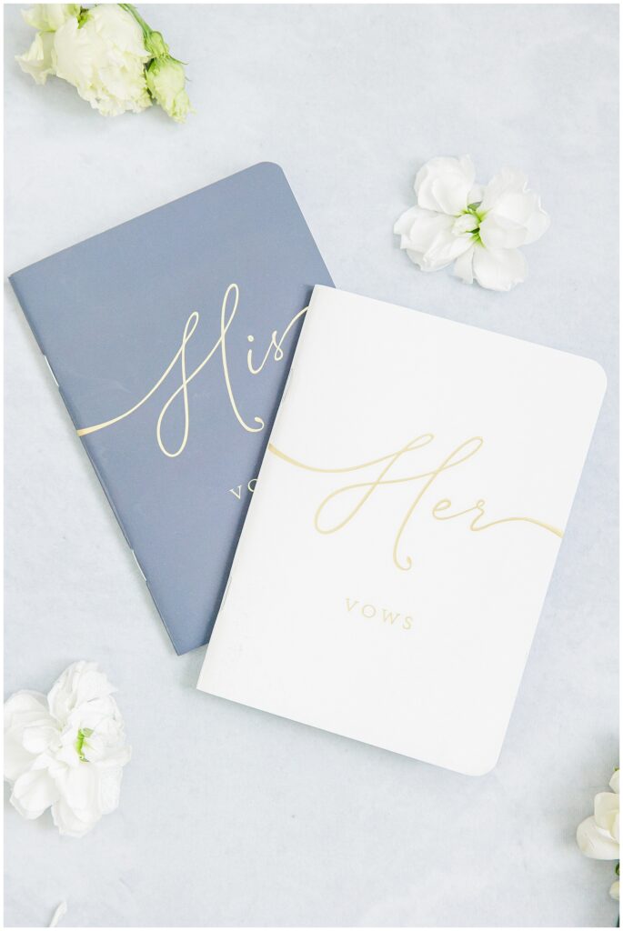 Two wedding vow booklets — one navy with “His Vows” and one white with “Her Vows” in gold script — surrounded by white flowers on a light blue surface.