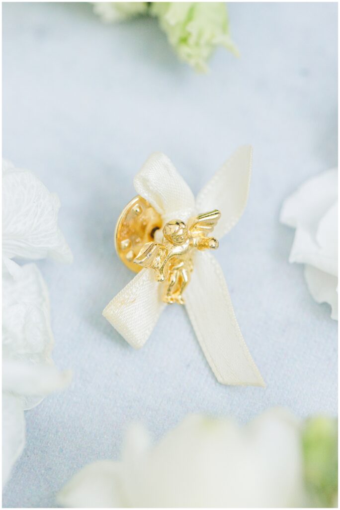 Small gold pin shaped like an angel with an ivory satin ribbon bow, displayed on a soft blue surface with white flower petals around it.